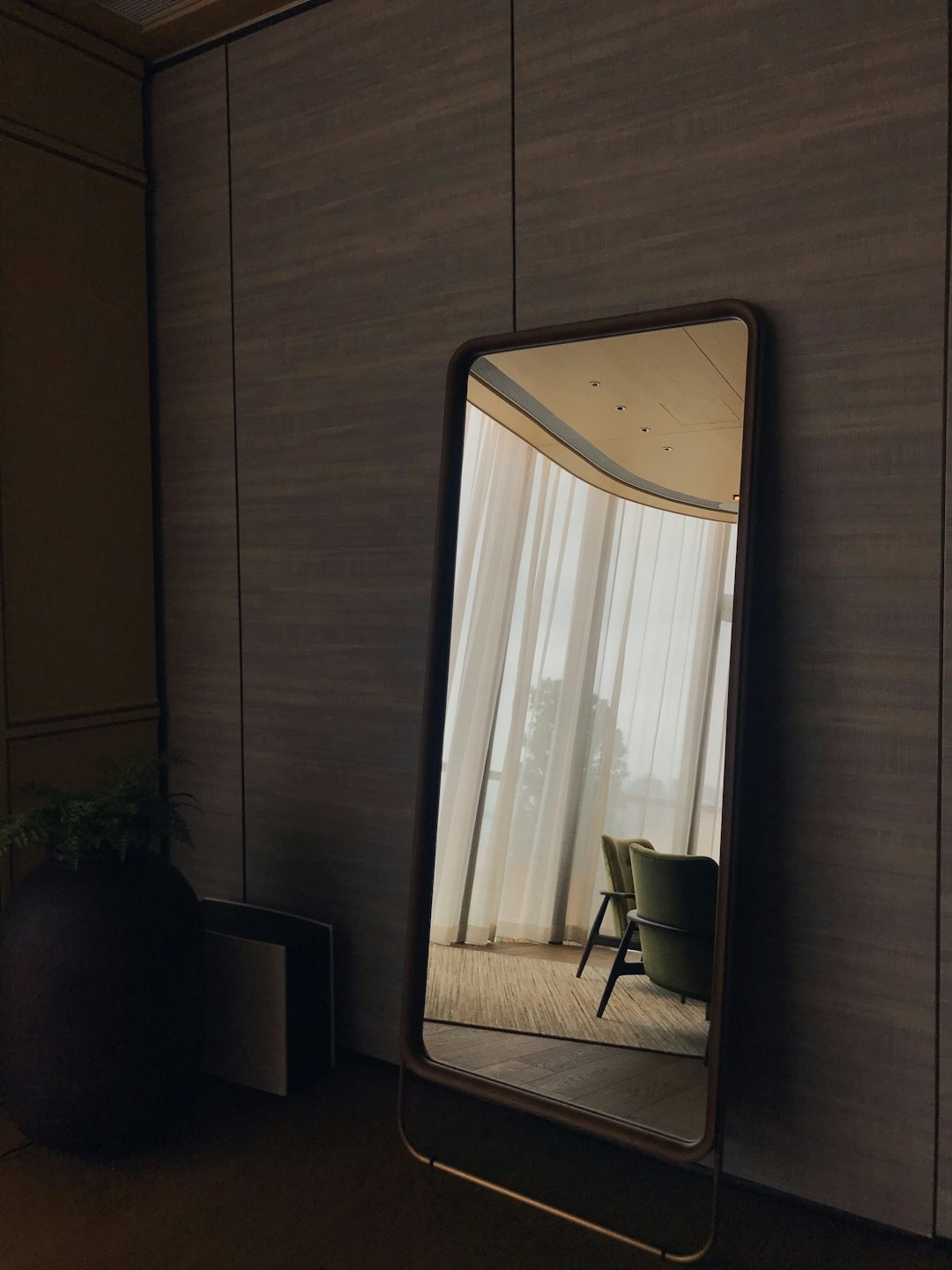 Modern floor mirror reflecting a bright room with armchairs.